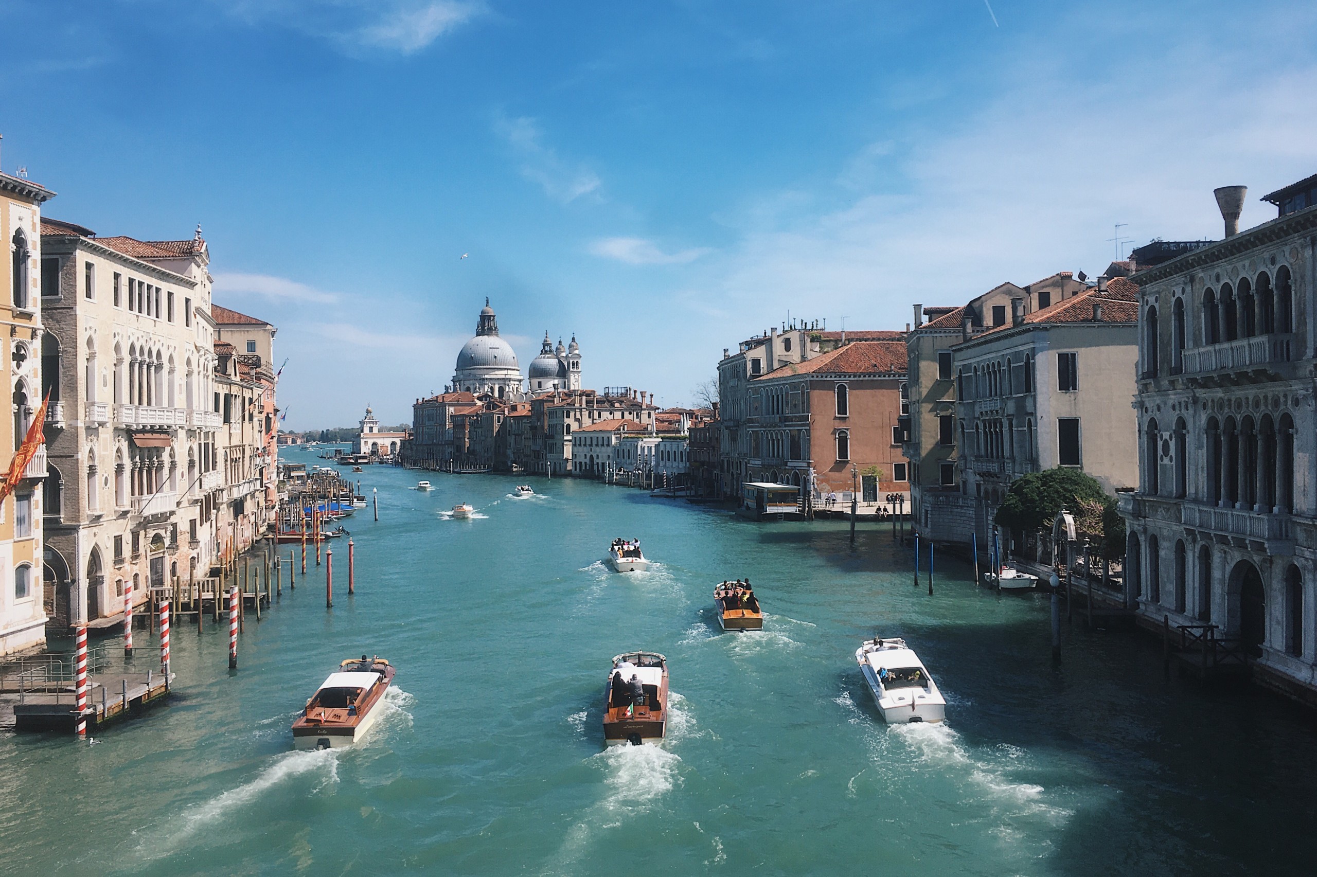 Ett stadslandskap från Venedig i färg. På bilden ser vi en bred å som rinner mot horisonten. På ån ser vi ett flertal båtar. Längs med ån står hus. Himlen är klarblå.