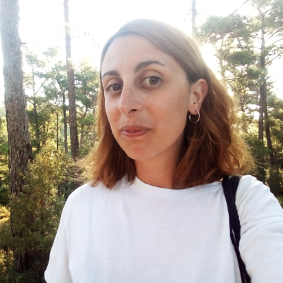 Caterina Avataneo is looking into the camera and smiling. She is in a forest and it's sunny. She is wearing a white t-shirt and has orange and brown shoulder-length hair.