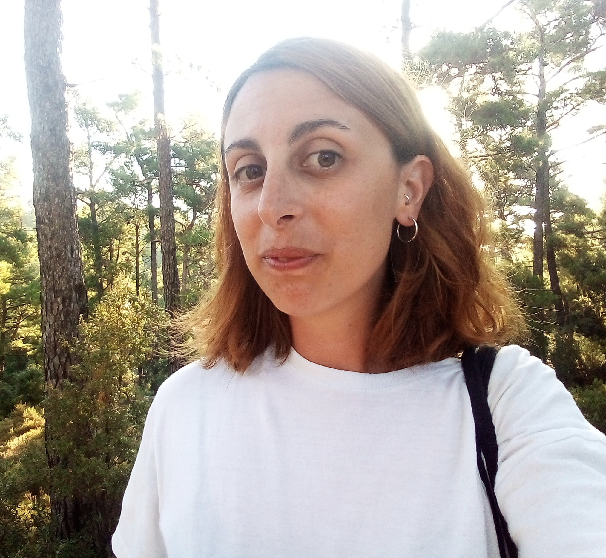 Caterina Avataneo is looking into the camera and smiling. She is in a forest and it's sunny. She is wearing a white t-shirt and has orange and brown shoulder-length hair.