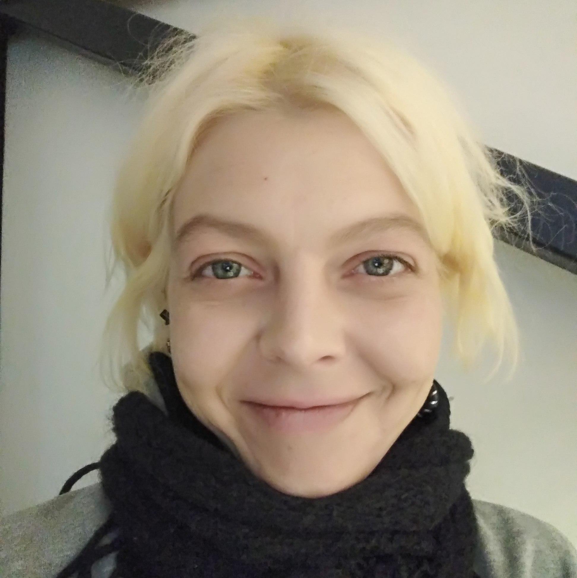 A portrait of Egle Nedzinskaite in colour. She has blonde straight hair. She is wearing a black scarf and is looking into the camera.