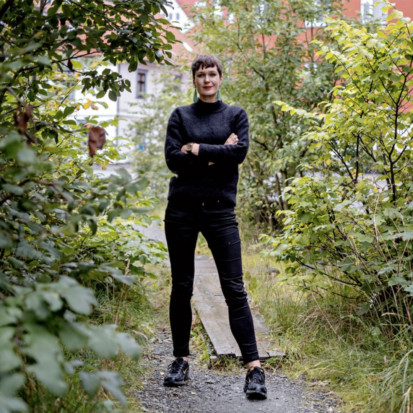 Eva Rowson is centered in the picture, where she is standing on a park path, surrounded by greenery. She is wearing black jeans and a black jumper. She is holding her arms crossed and is smiling.