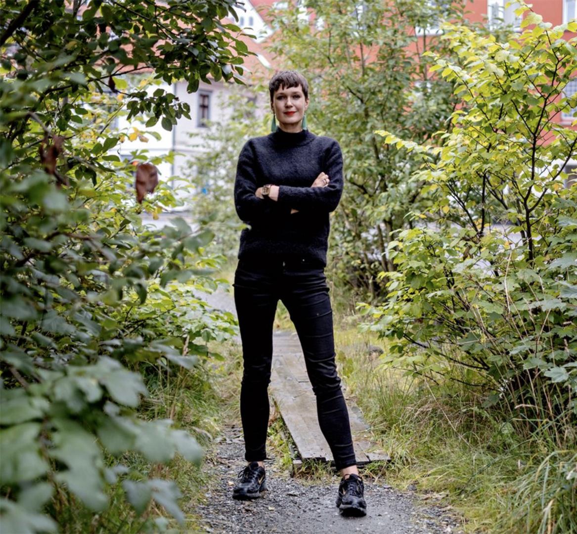 Eva Rowson is centered in the picture, where she is standing on a park path, surrounded by greenery. She is wearing black jeans and a black jumper. She is holding her arms crossed and is smiling.