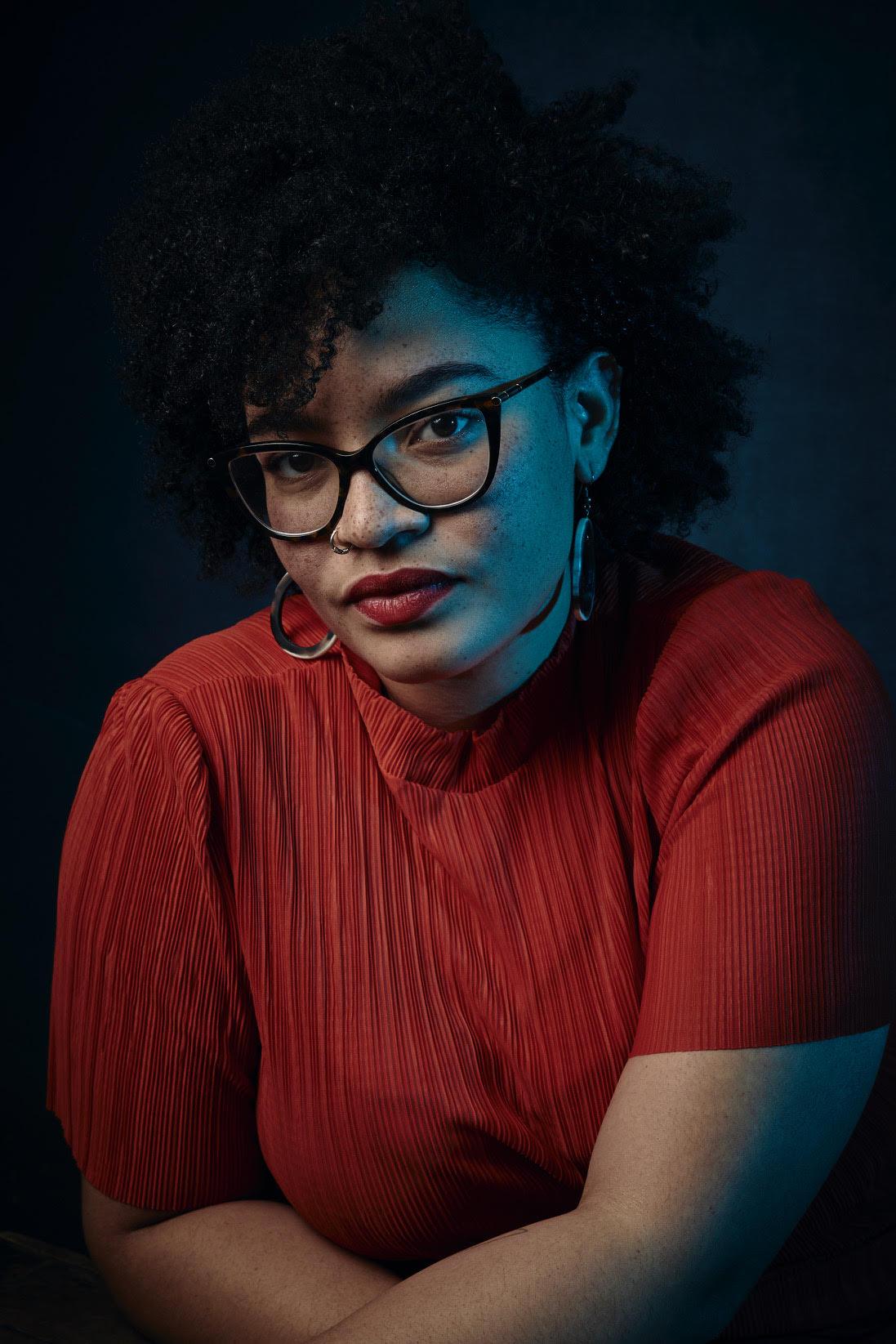 A person with a brown skin and dark curly hair wearing black glasses and a red shirt.