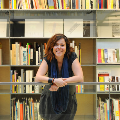 Gemma is leaning on a railing and looking into the camera smiling. She is wearing a grey dress and blue scarf and has dark hair with orange highlights. She is in front of a book shelf.