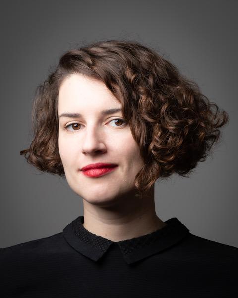 Miriam Wistreich, a white-skinned person with short dark hair is standing in front of a grey backdrop looking at the camera. Miriam has a black shirt on and red lipstick.