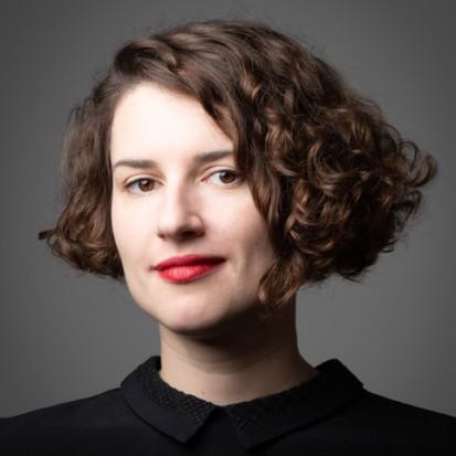 A studio portrait Miriam Wistreich in colour. She has short, brown curly hair and red lipstick. She is wearing a black collared shirt and is looking into the camera.