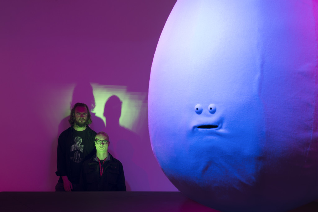 Taiteilijat Erkka Nissinen ja Nathaniel Mellors katsovat suurta puhuvaa munanmuotoista fiiguuria, joka on osa heidän installaatiota.