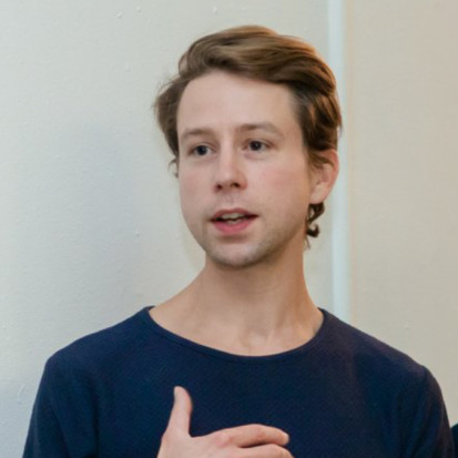 Yates Norton is centered in the image, which is taken indoors. He is talking and is not looking into the camera. He has short, light brown hair and he is wearing a dark blue t-shirt.