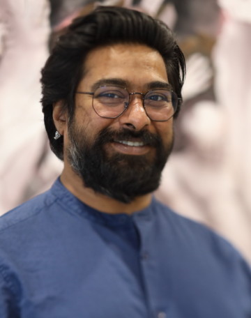 Half-portrait of Ali Akbar Mehta wearing round glasses and blue shirt with black hair and black beard. Ali looks straight into camera, smiling.