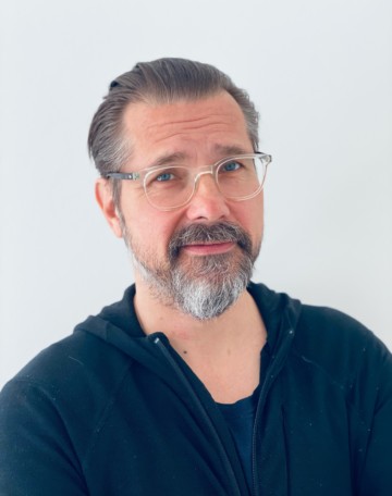 Half-portrait of Esa Alanne. He is looking straight to the camera. He is wearing glasses and blue shirt and has a beard.