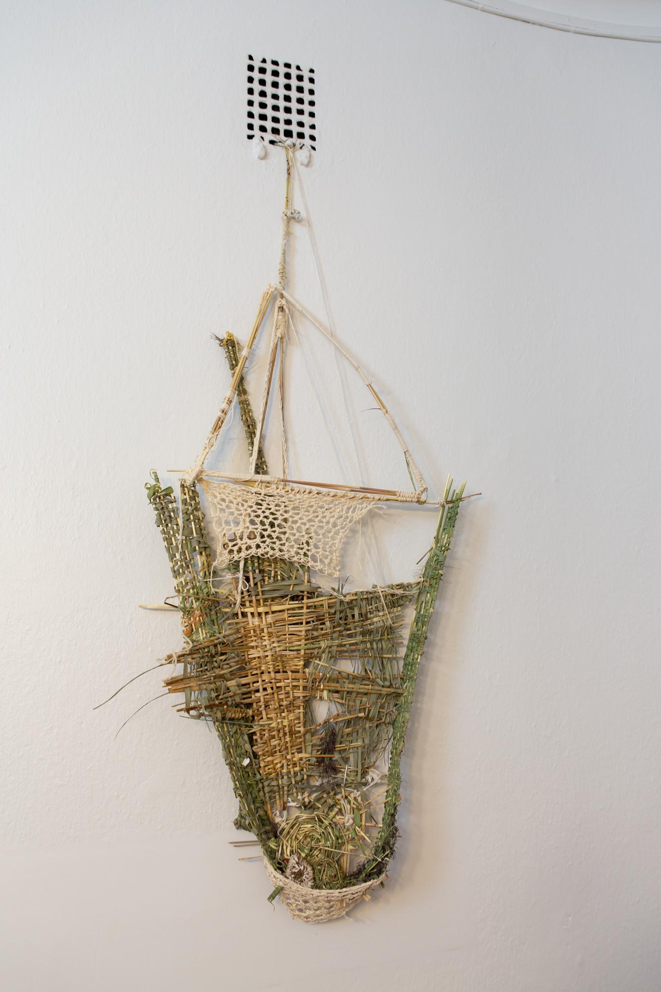 Artwork Seven Recipients at the frame office, consisting of sticks and strings woven together to form a roughly shaped triangular basket-like object, is hung on a white wall.