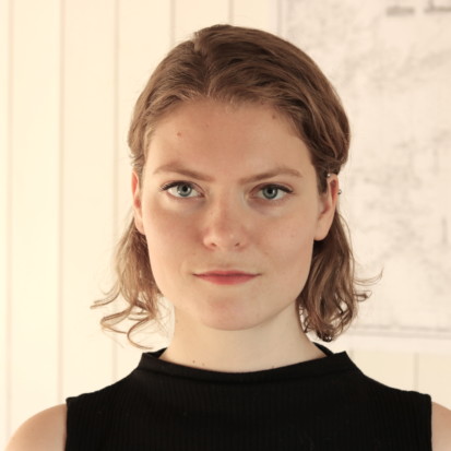 Ann Mirjam Vaikla, a white-skinned person with red lipstick and shoulder-length, curly blonde hair. Framed from shoulders up Ann Mirjam is standing in front of a white backdrop.