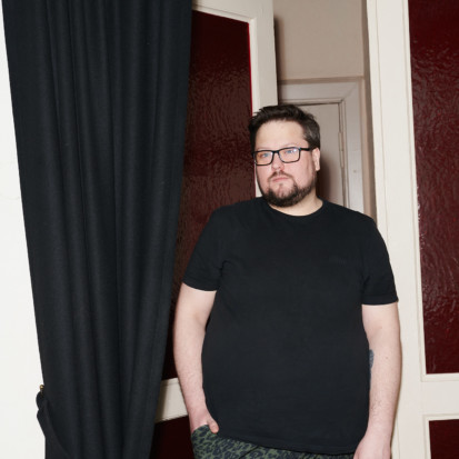 Edvinas Grinkevičius, a white-skinned person with dark brown short hair, dark brown beard, eyeglasses. Framed from knees up, Edvinas is wearing a black t-shirt, dark jeans and is leaning towards a door opening. A black curtain is on the left of Edvinas.