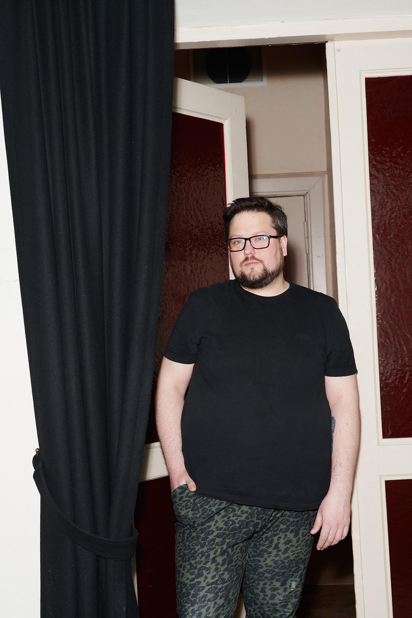 Edvinas Grinkevičius, a white-skinned person with dark brown short hair, dark brown beard, eyeglasses. Framed from knees up, Edvinas is wearing a black t-shirt, dark jeans and is leaning towards a door opening. A black curtain is on the left of Edvinas.