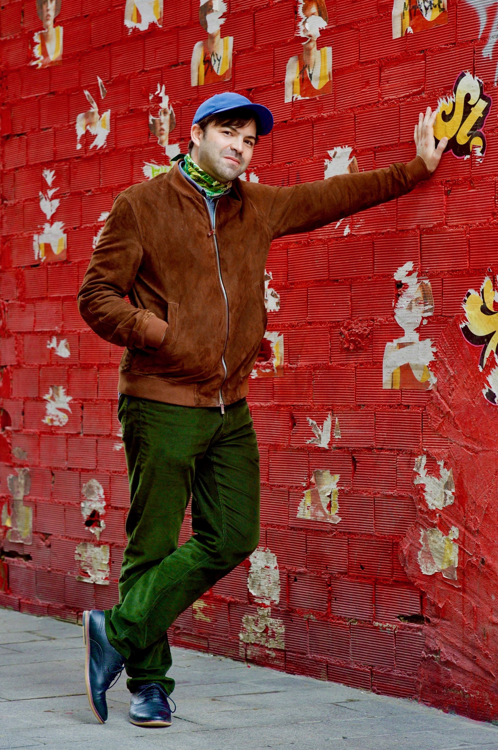 Portrait of Francisco Martínez. Francisco has short dark hair, green trousers, brown jacket, a colorful scarf and a blue cap on. Francisco is leaning with his left hand against a red wall.