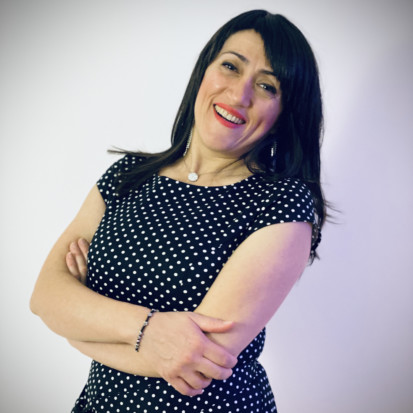 Portrait of Abir Boukhari. Abir has a long black hair and a black dress with white dots. Abir is smiling while crossing her hands. The background is white.