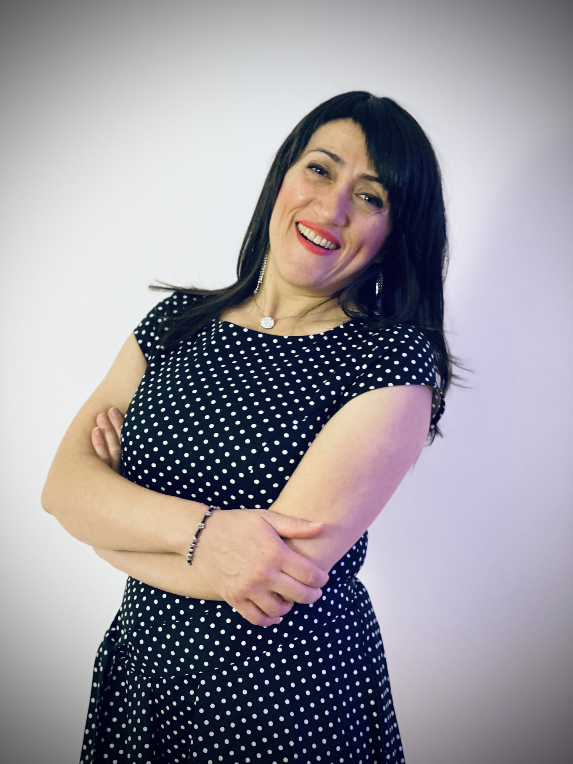 Portrait of Abir Boukhari. Abir has a long black hair and a black dress with white dots. Abir is smiling while crossing her hands. The background is white.