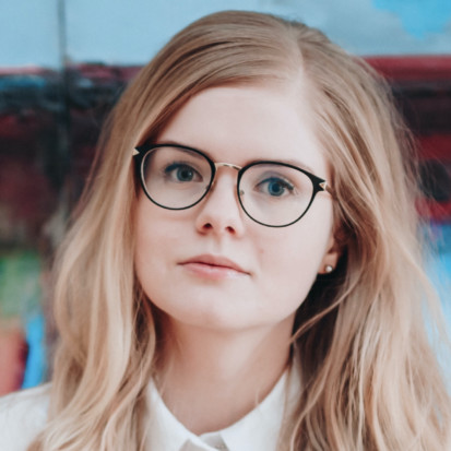 Portrait of Joanna Hoffmann. Joanna has long blonde hair and black eyeglasses on. Joanna is wearing a white shirt. The background is blue and red.