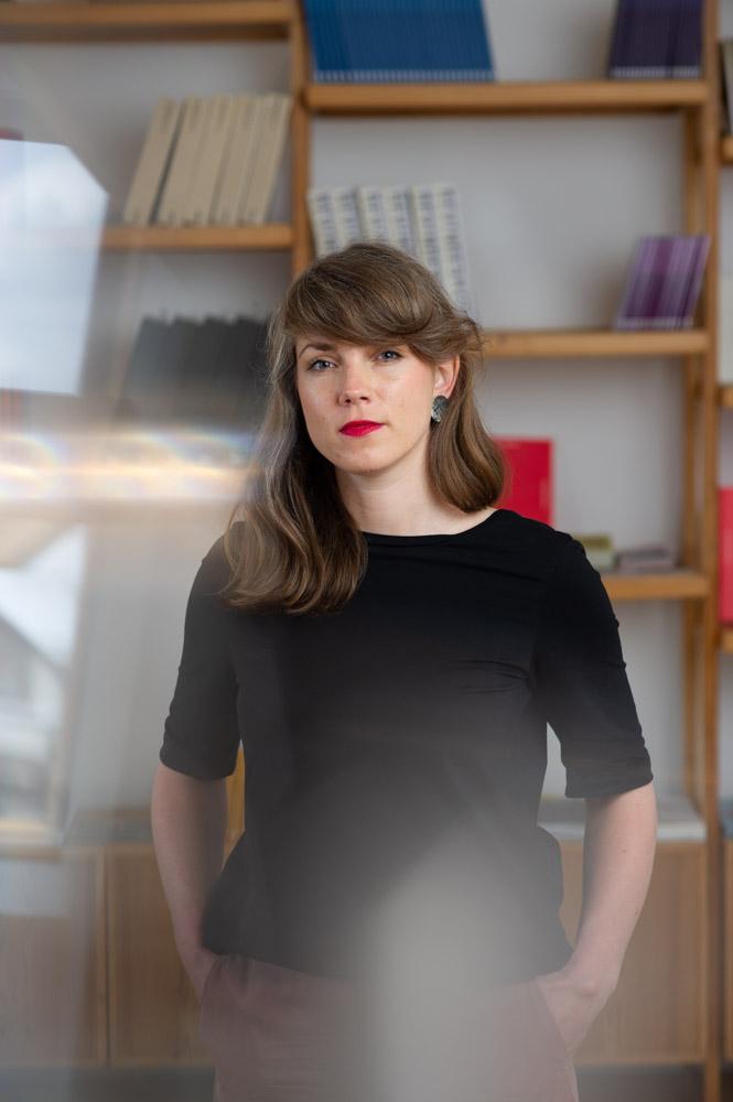 Portrait of Andra Silapētere. Andra has long dark brown hair, red lipstick and a black shirt on. Andra is standing in front of a bookshelf with her hands in her pockets.