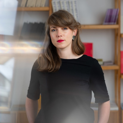 Andra Silapētere, a white-skinned person with a brown long hair, forelock and red lipstick. Framed from lower waist up Andra is standing in front of a bookshelf with her hands in her pockets.