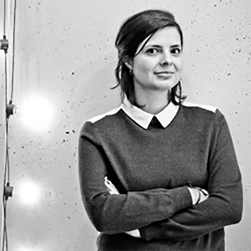 Portrait in black and white of Mechu Rapela. Mechu has dark shoulder length hair and a sweater on. Mechu is standing with her hands crossed in front of a wall.