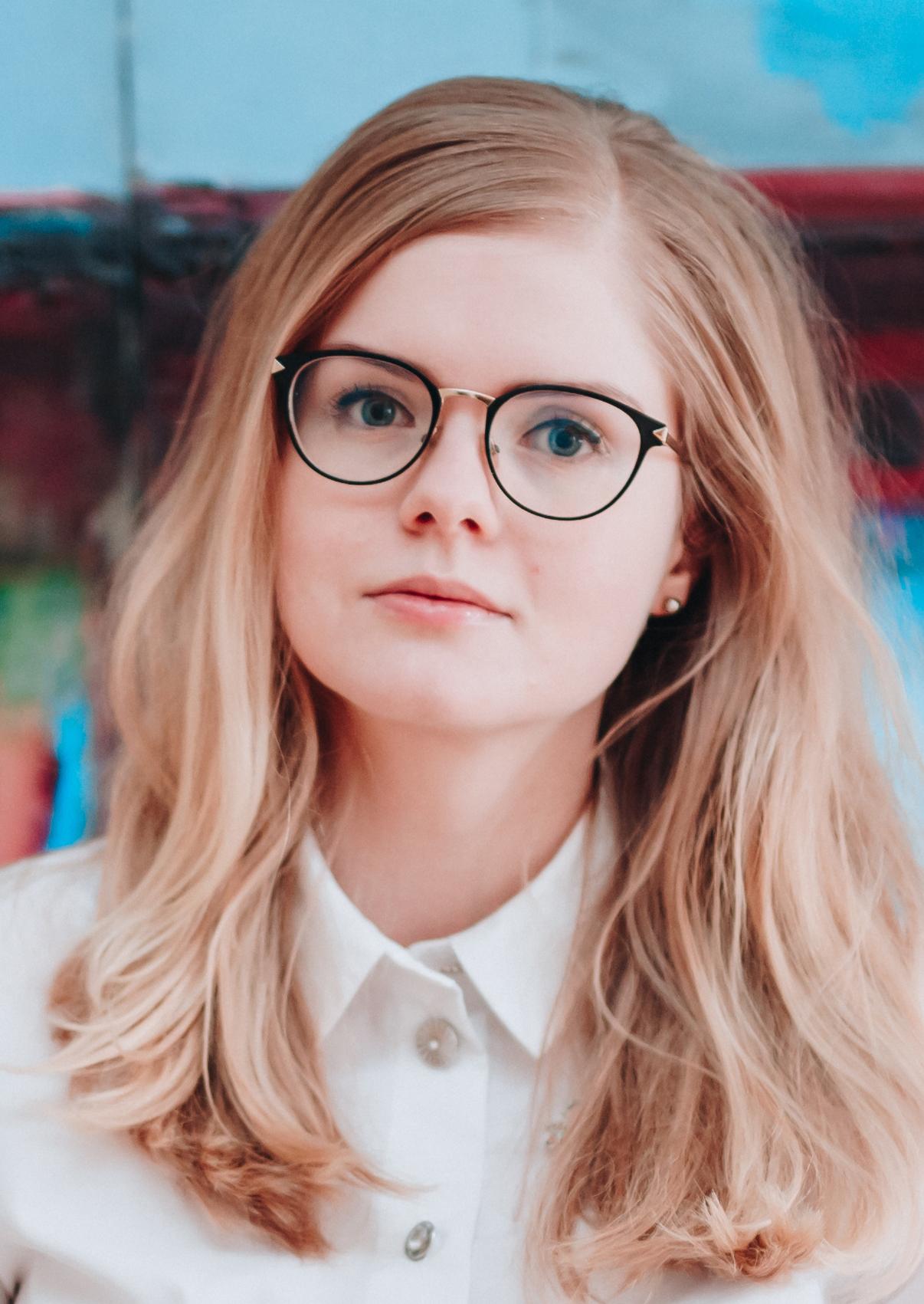 Joanna Hoffmann, a white-skinned person with long blonde hair and black eyeglasses. Joanna is wearing a white shirt with buttons. Framed from chest up, Joanna is standing in front of a blue and red backdrop.