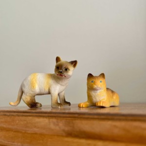 There are two cat figurines on a brown table facing the viewer. The cat figurines are against light background and another of them is standing and the other is laying down.