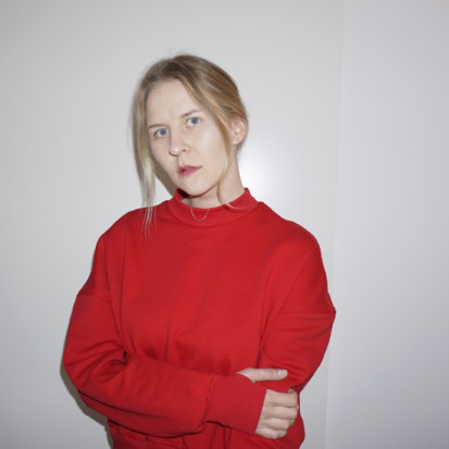 Lilian Hiob, a white-skinned person with blonde hair and red lipstick. Framed from waist up, Lilian is wearing a bright red sweater. Lilian has crossed her hands and is standing in front of a white backdrop.