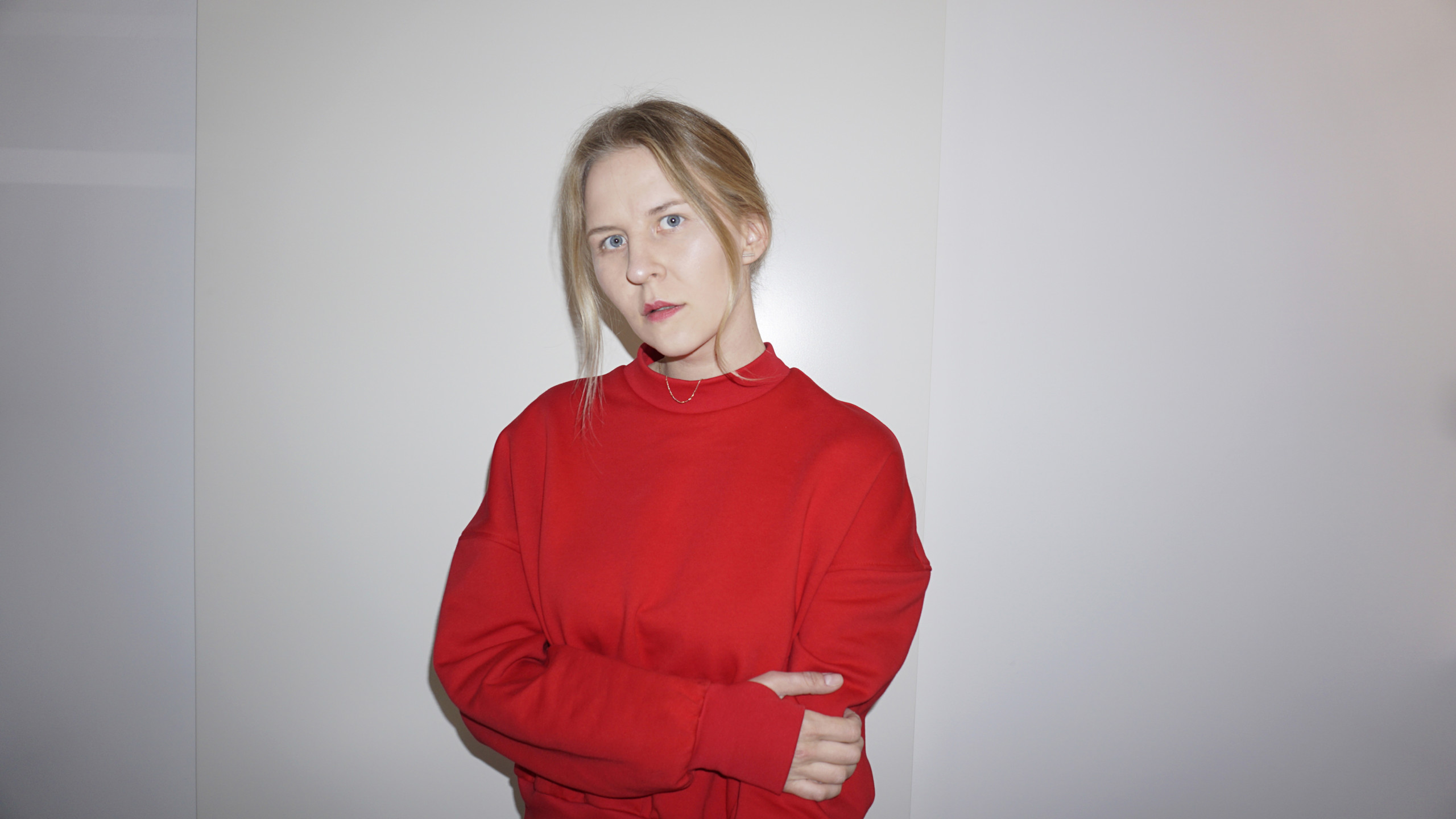 Lilian Hiob, a white-skinned person with blonde hair and red lipstick. Framed from waist up, Lilian is wearing a bright red sweater. Lilian has crossed her hands and is standing in front of a white backdrop.