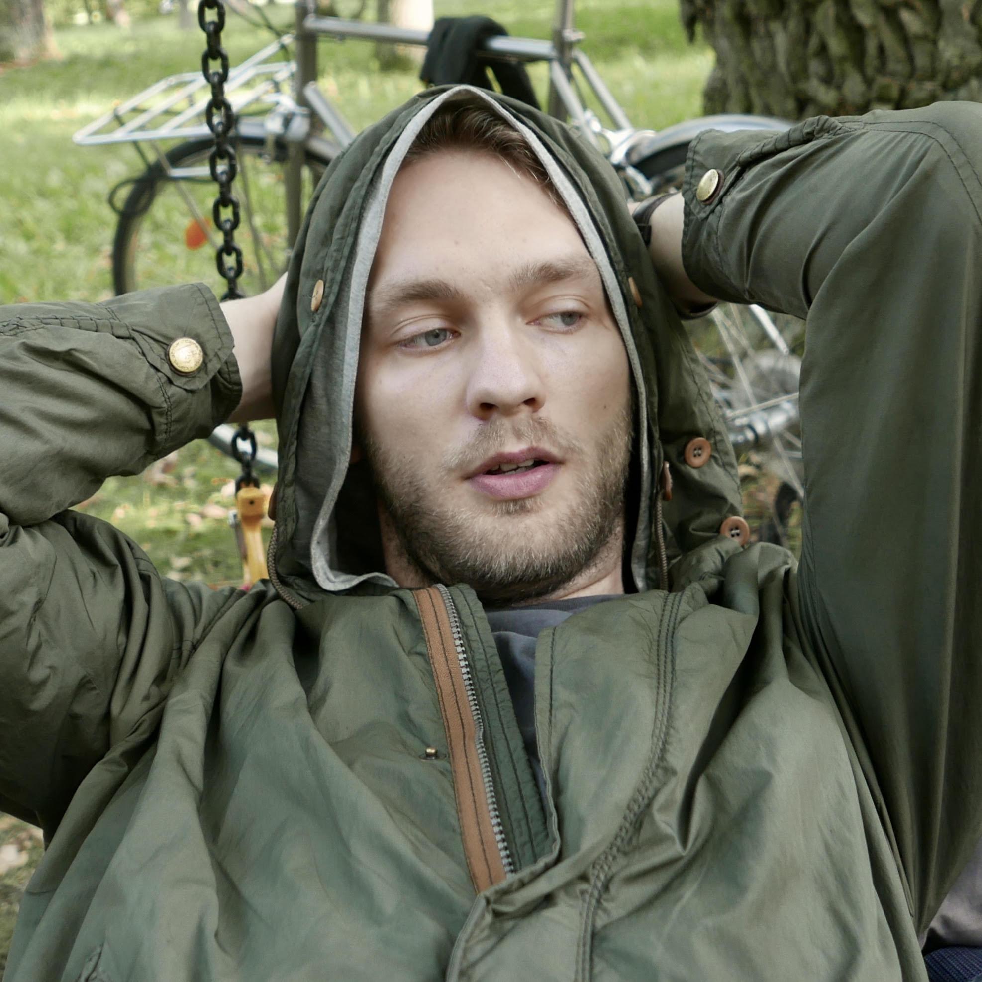 Marten Esko, a white-skinned person with blonde hair and blonde beard and mustache. Marten is wearing a green jacket with a hoodie. Framed from the waist up, Marten has placed his hands behind his head in the picture. The backdrop of the picture features a tree, grass and a bicycle.