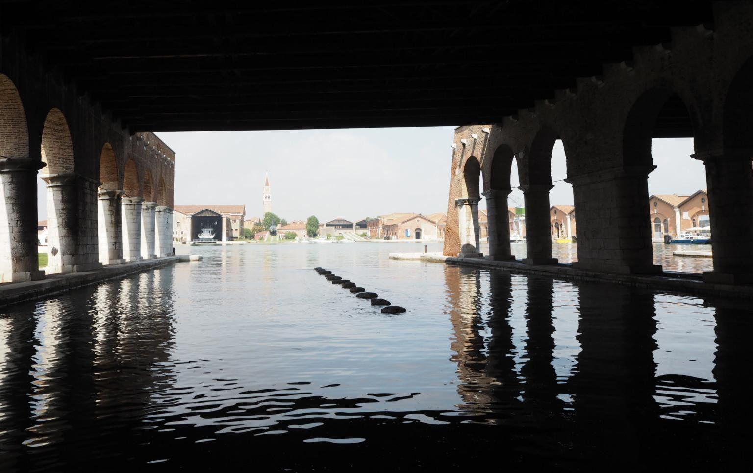 En bild i färg av utställningsområdet och gamla skeppsvarvet Arsenale i Venedig. På bilden kan vi se vatten som täcker nedre delen av bilden. På vänster och höger sida av bilden sträcker sig stora mörka pelare upp mot himlen. 