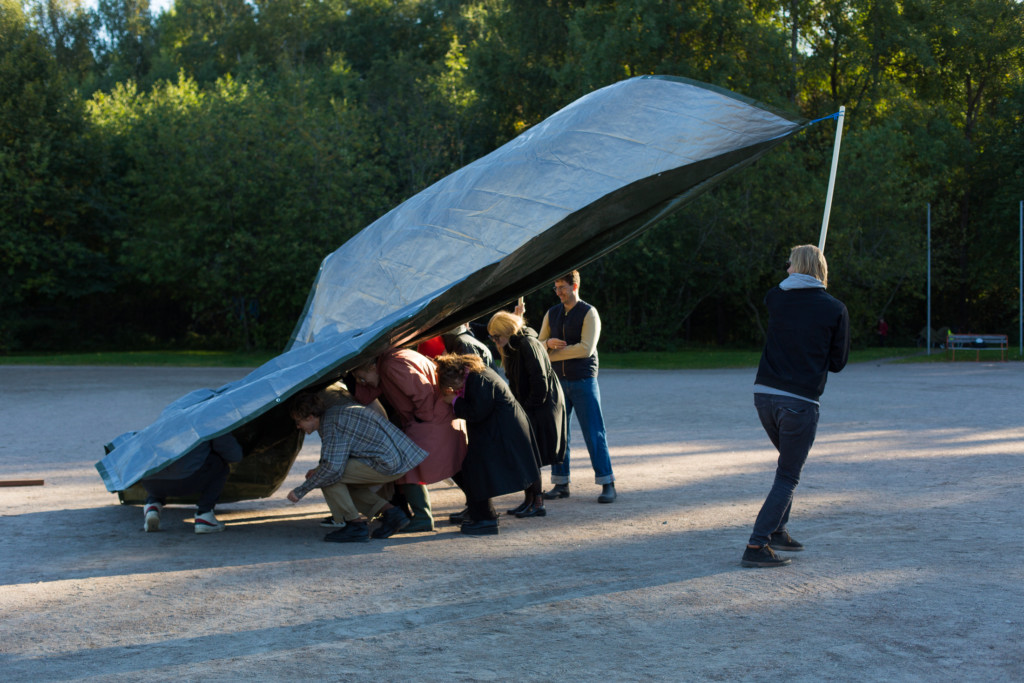 Medium long-shot of a group of people hunching down together while a person throws a big tarp over them. Atmosphere is playful.