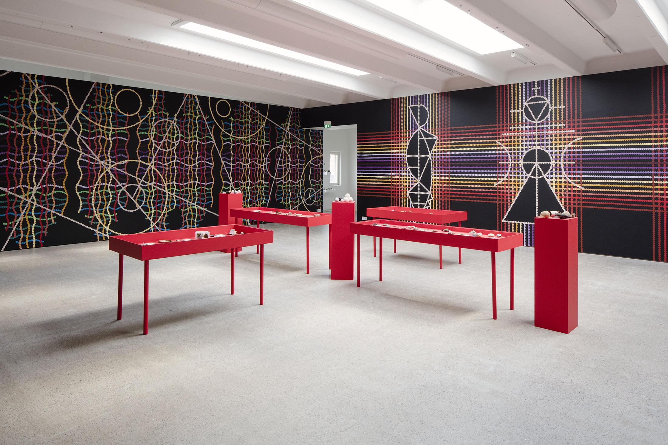 A vast exhibition space, with four red tables and three red pedestals under bright white light. There are vibrant dizzying patterns on the wall.