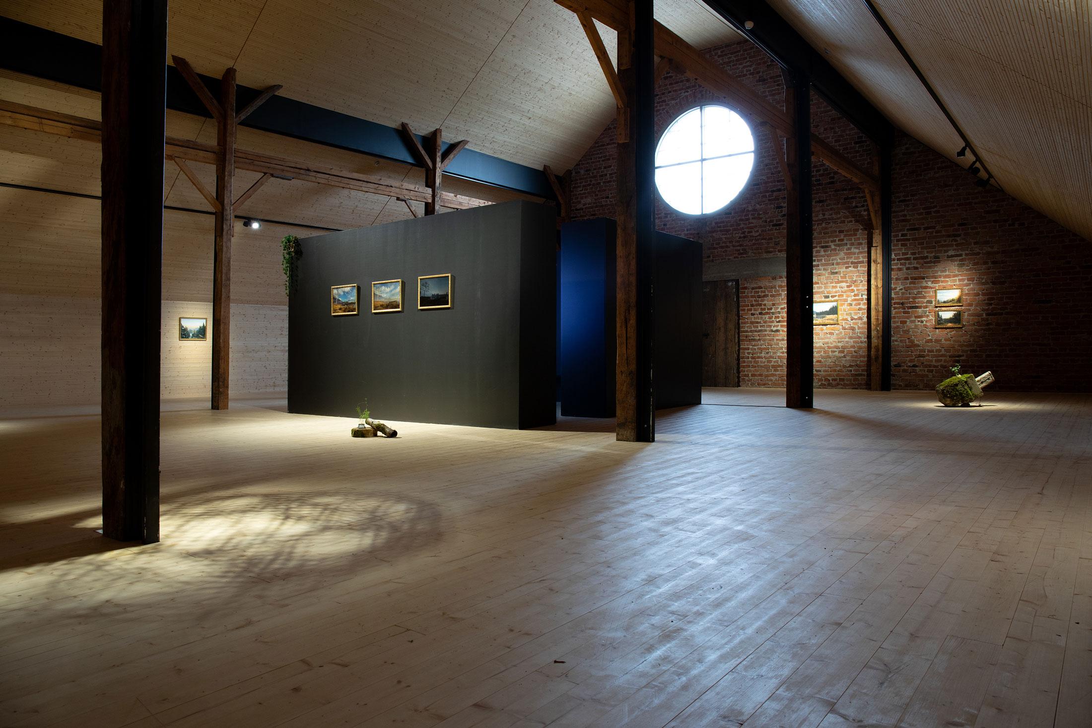 A vast, dimly lit, industrial looking exhibition space. Some artworks like framed images, and sculptures are minimally spread out and distantly visible.