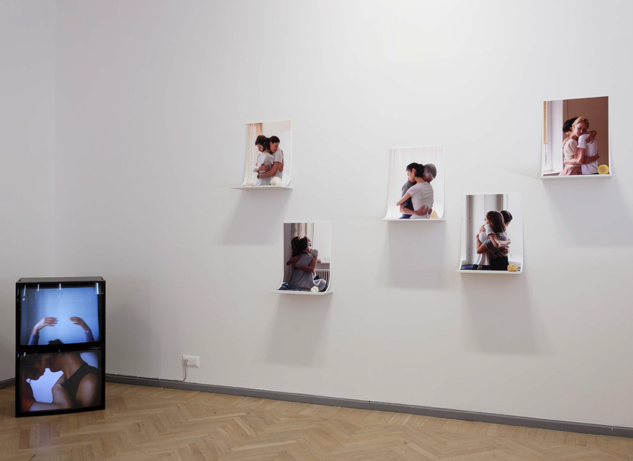 Exhibition space with photographs of hugging people on the walls. On the floor there is screen with video or image on it.