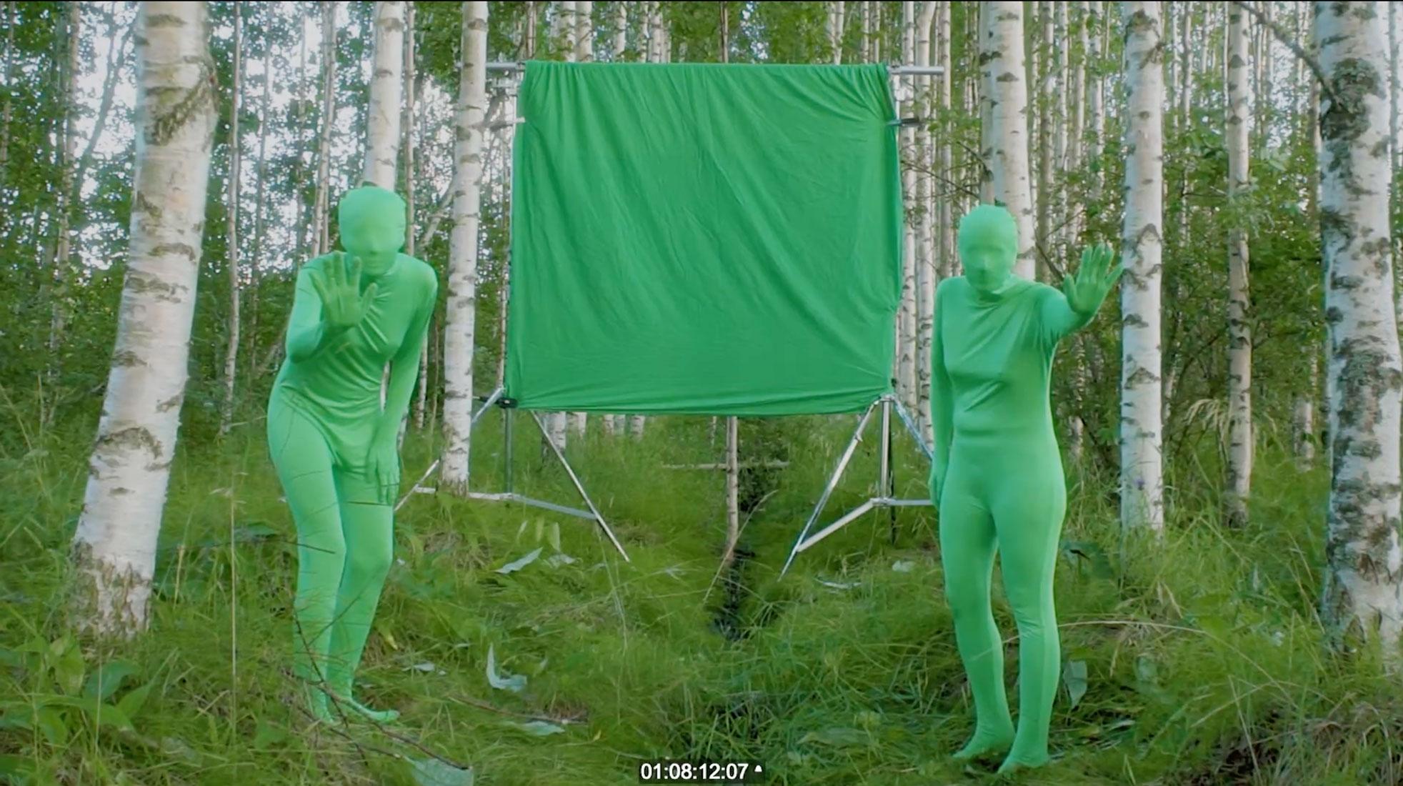 Two humans, covered in green fabric from head to toe, stand in a forest area with green grass and birch trees. There is a green fabric screen hanging in between them, and they are both looking into the camera.