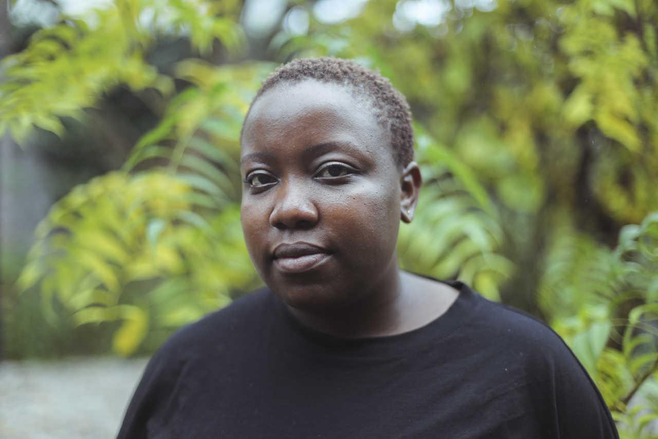 Yolande Zola Zoli van der Heide, a black-skinned person with short brown hair stands in front of a backdrop with plants looking into the camera. Framed from the shoulders up, van der Heide is wearing a black shirt.
