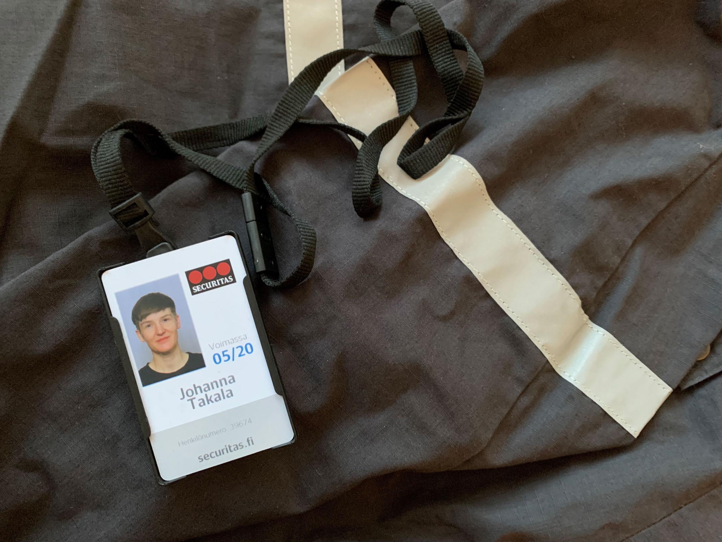 Black uniform-like fabric, with a security company personnel's identity card kept on it. The card carries artist Pilvi Takala's headshot with the name Johanna Takala under it, and a logo of the company Securitas.
