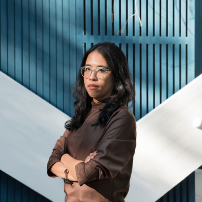 Curator Christina Li, with dark wavy hair and tawny skin, is pictured against a blue wooden wall of the Finnish pavilion at the Venice Bienale. She is looking straight into the camera, wearing round glasses, with her arms crossed.