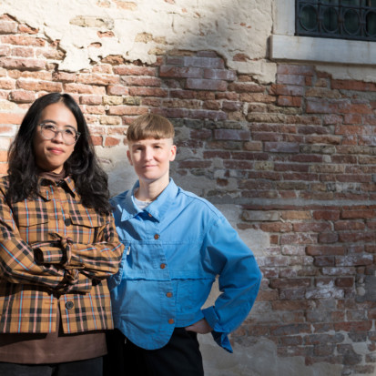 Artist Pilvi Takala, with white skin and short ash-blonde hair is pictured standing next to Curator Christina Li, with dark wavy hair and tawny skin. They are both looking into the camera, standing in front of a distressed brick wall on a bright sunny day.