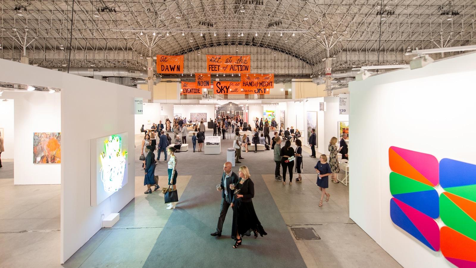 A vast open exhibition space of EXPO CHICAGO, the international exposition of contemporary modern art. The space, which looks like an aircraft hangar, is filled with visitors engaging with the art.