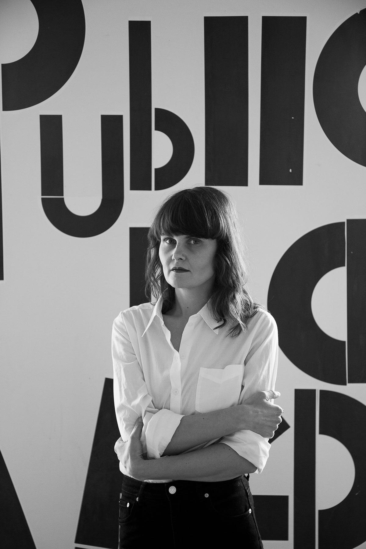 A black and white photo, Eliisa directly looking into the camera with a slightly serious expression on their face, a phrase 'Publics is a verb' by Kathrin Böhm is pasted on the wall behind