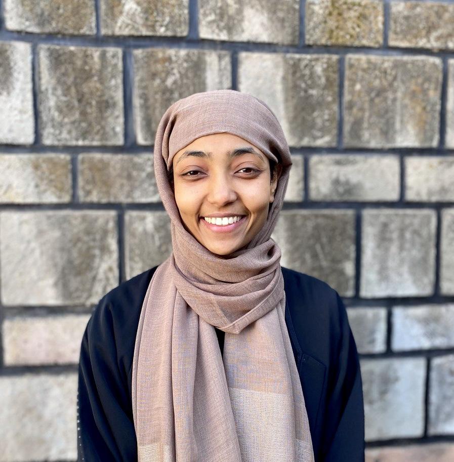 A headshot of sarah, a black-skinned person, in front of a cinder block wall. she's smiling and looking at the camera. she's wearing a light brown headscarf and black shirt.