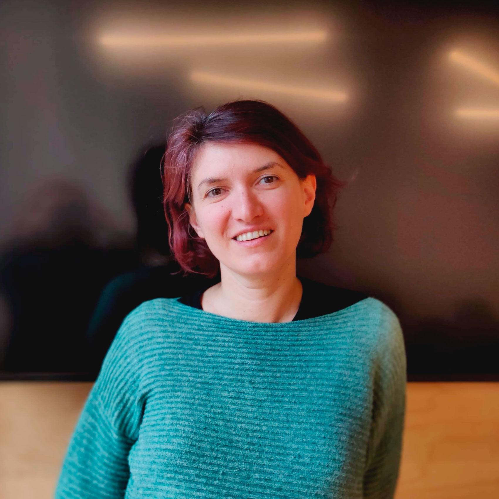 Raluca Voinea, a white-skinned person with dark hair, looks into the camera with a smile. She is is wearing a bright sweater, against a black background.