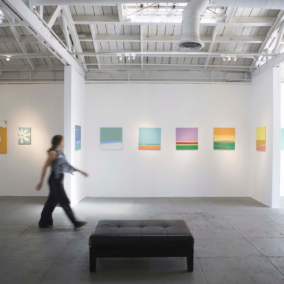 A person walking through white walled gallery space. On the wall there is colourfull small paintings.