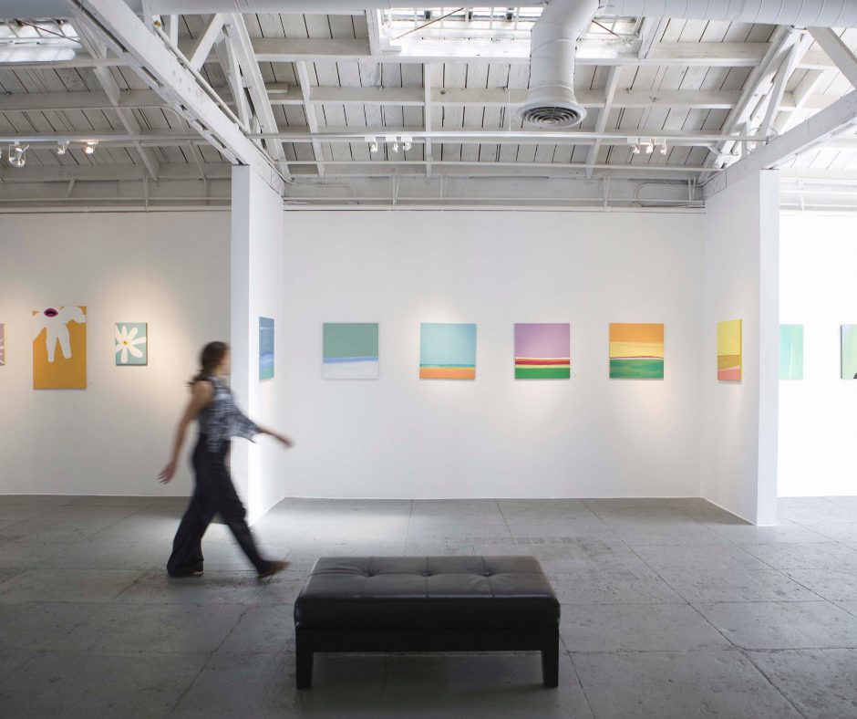 A person walking through white walled gallery space. On the wall there is colourfull small paintings.