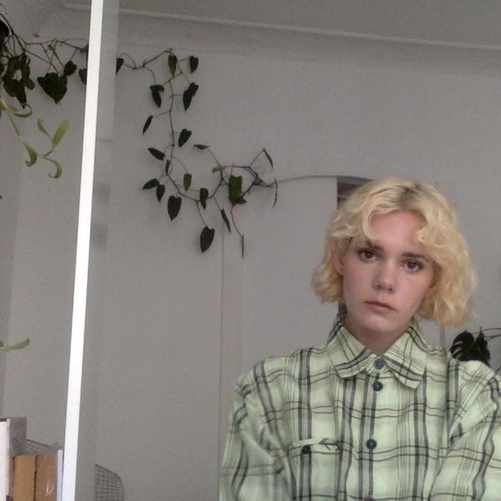 Christina Gigliotti, a white-skinned person with blonde short hair is standing in front of a white backdrop looking into the camera. Gigliotti has a plaid green blouse shirt on.