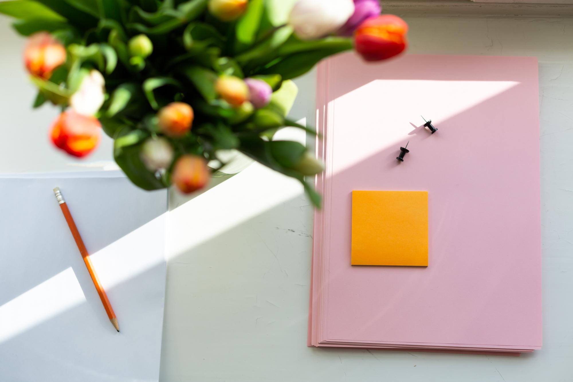 Pink paper, orange post-its, pencil and tulip on an office desk.