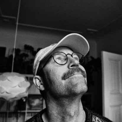 Leif Magne Tangen, a white-skinned person with short hair and moustache is looking up and smiling. The image is black and white and the backdrop of the image is displaying a room, a book shelf and an open door. Framed from the shoulders up, Leif is wearing a t-shirt, round glasses and a cap.