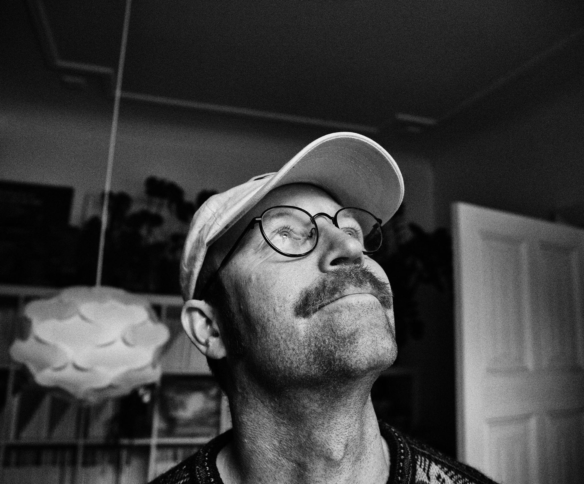 Leif Magne Tangen, a white-skinned person with short hair and moustache is looking up and smiling. The image is black and white and the backdrop of the image is displaying a room, a book shelf and an open door. Framed from the shoulders up, Leif is wearing a t-shirt, round glasses and a cap.